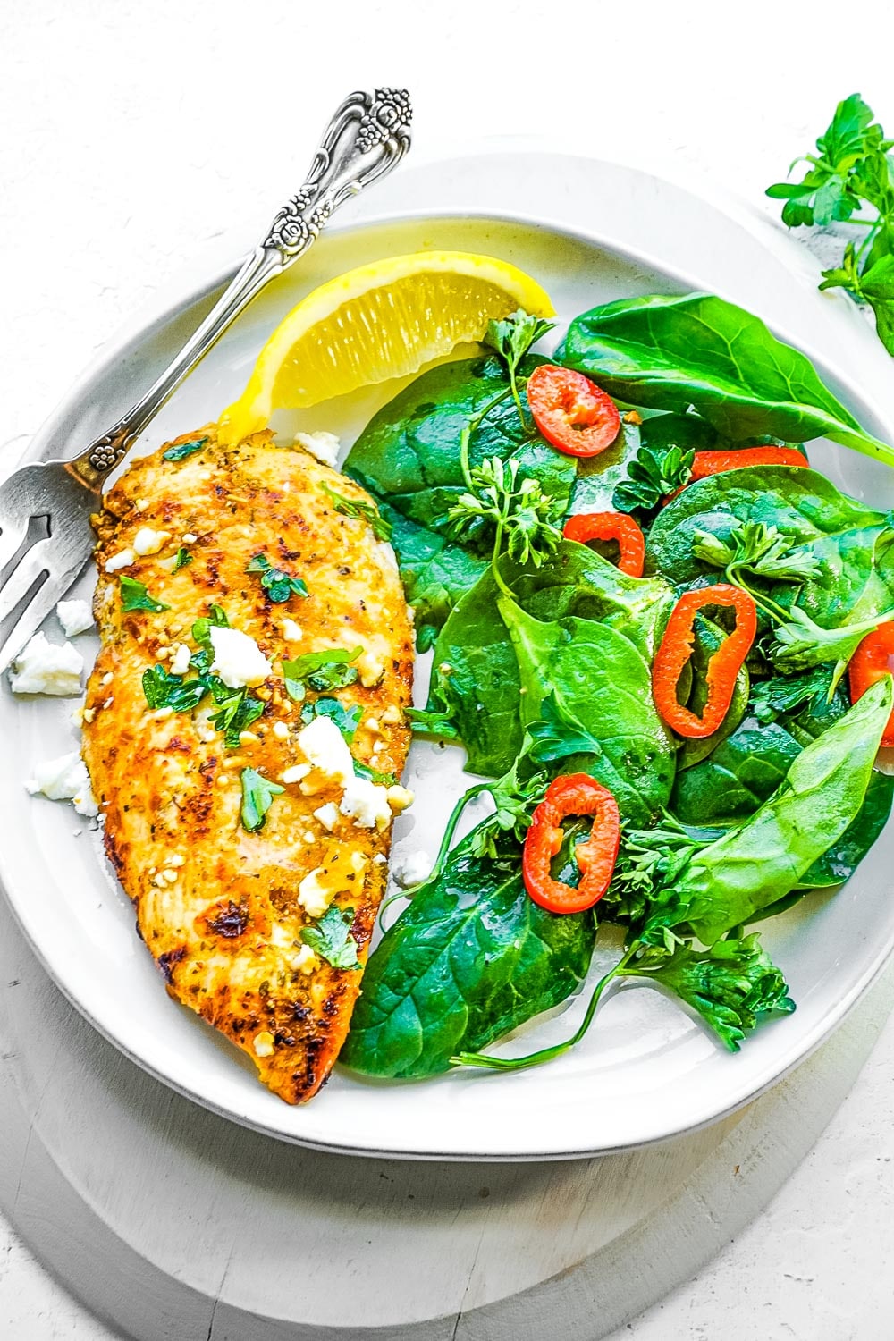 A piece of Greek chicken on a plate with a spinach salad and a lemon wedge.