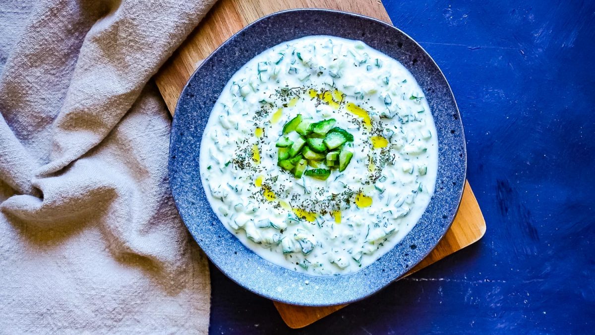 An overhead picture of cucumber yogurt sauce on a dark bowl garnished with mint and olive oil.