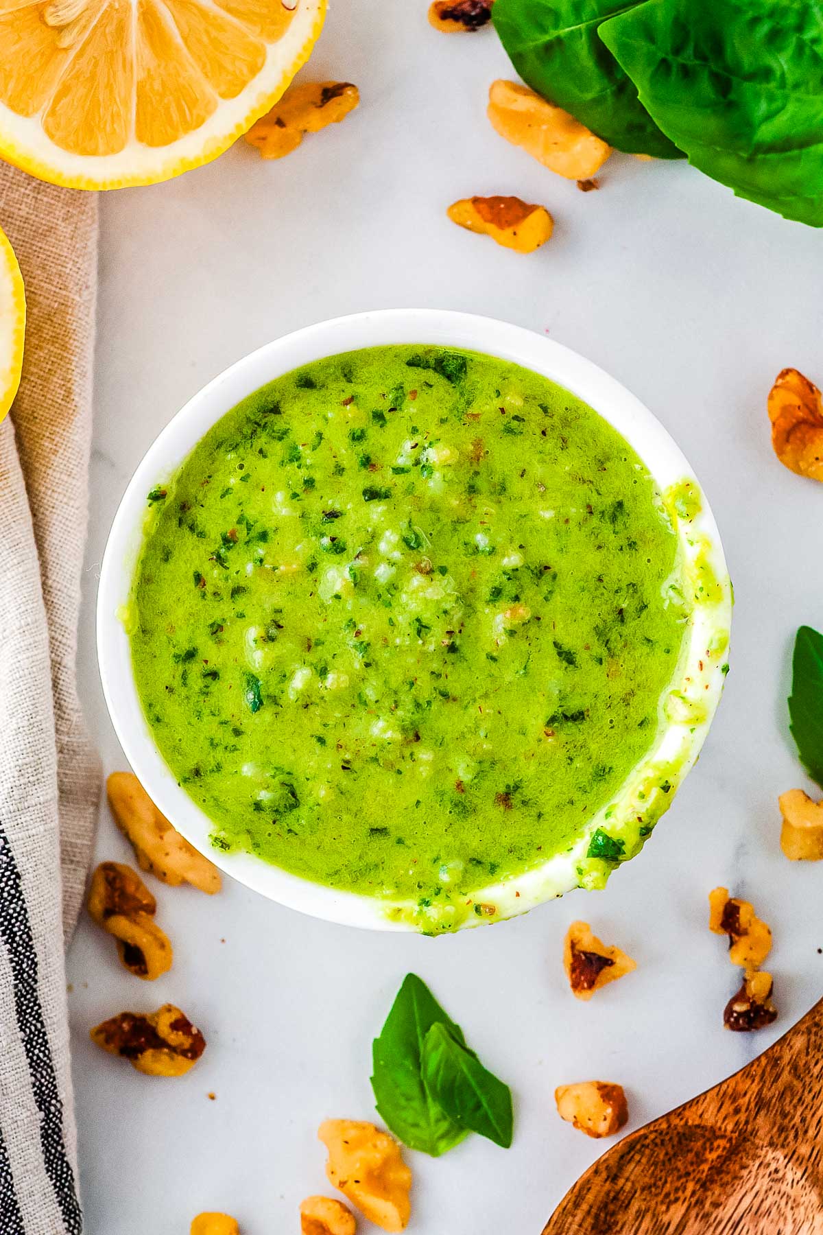Overhead view of walnut basil pesto in a white ramekin surrounded by fresh basil leaves, walnuts, lemon slices, and a wooden spoon.