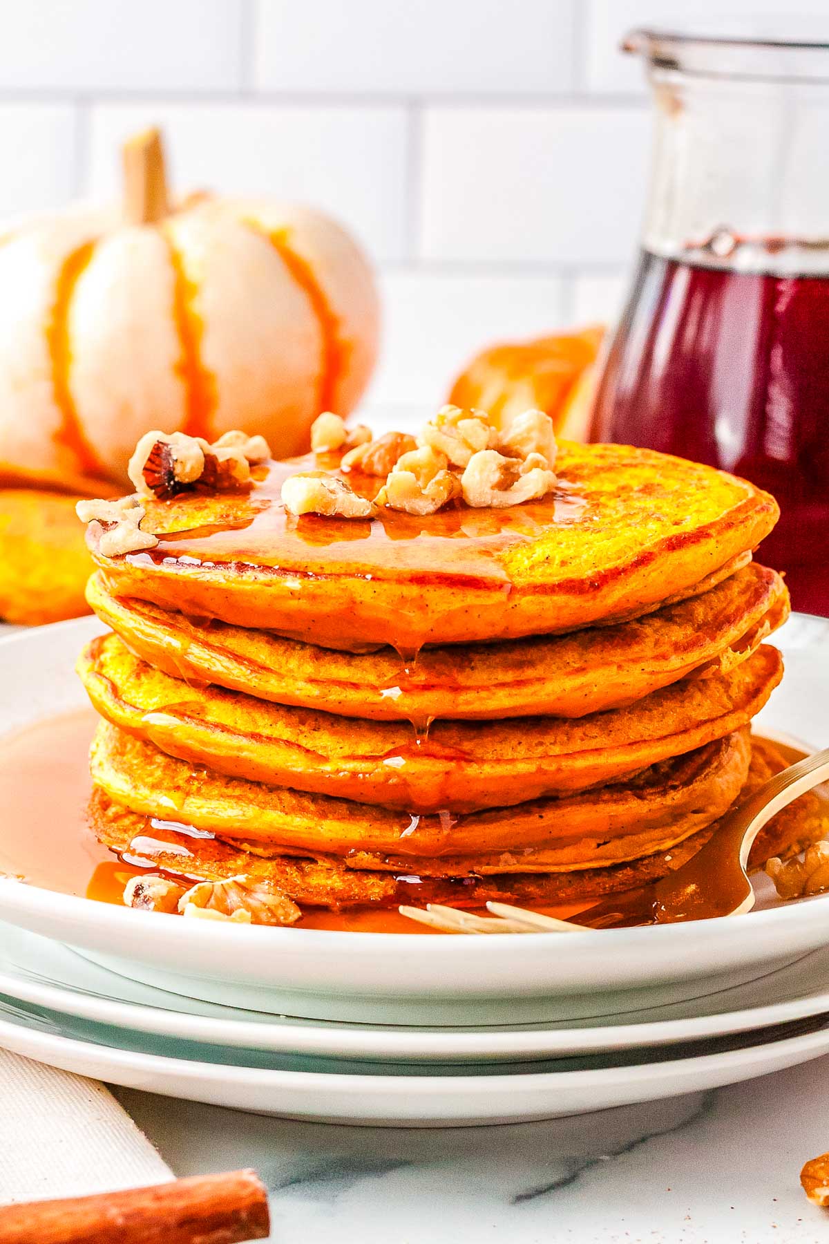 Tall stack of pumpkin cottage cheese pancakes drizzled with maple syrup and sprinkled with walnuts, with pumpkins in the background.