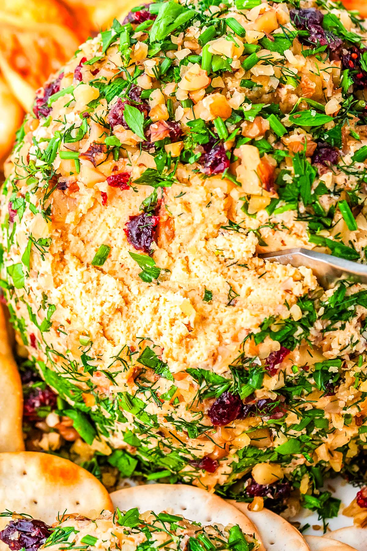 Close-up of vegan cheese ball showing creamy interior and coating of fresh herbs, cranberries, and toasted nuts.
