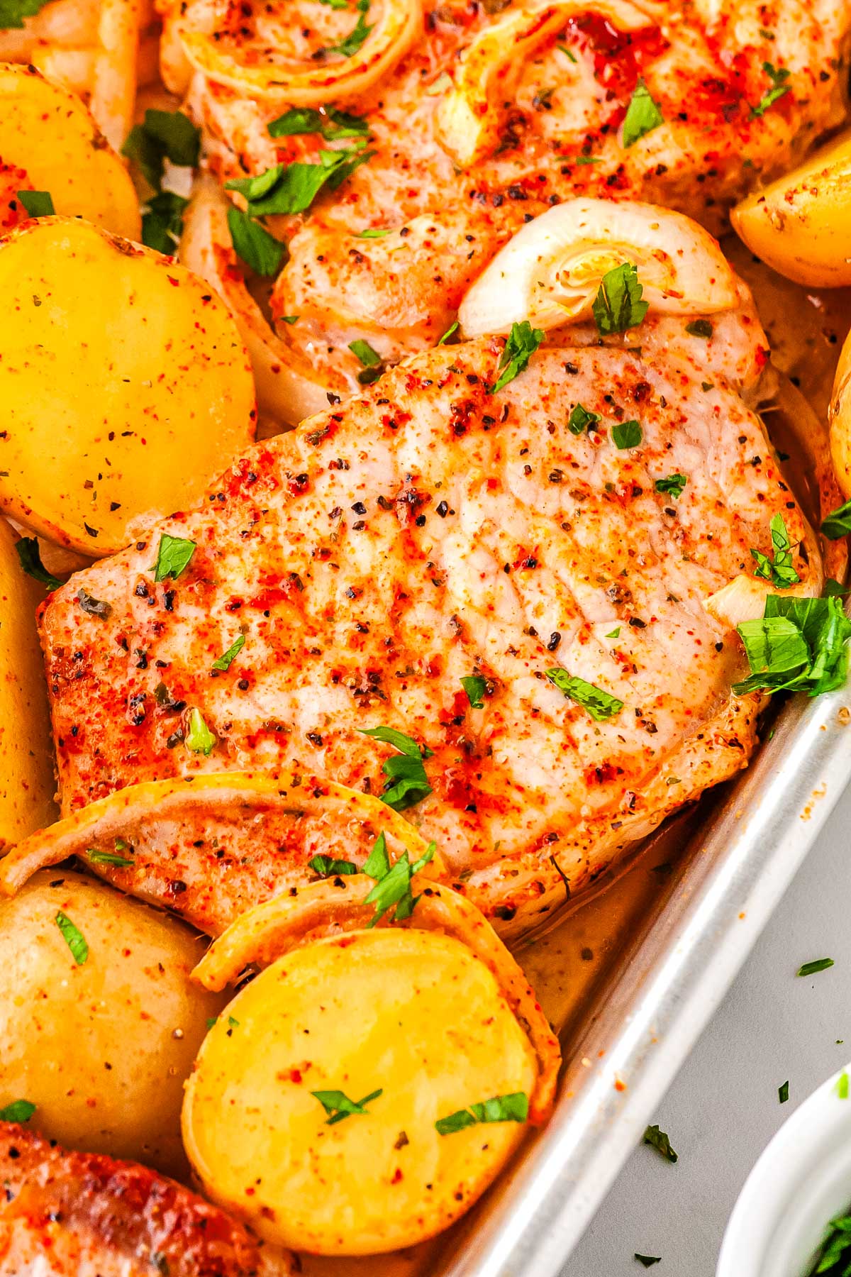 Close-up of baked pork chop surrounded by roasted potatoes and onions, sprinkled with parsley.