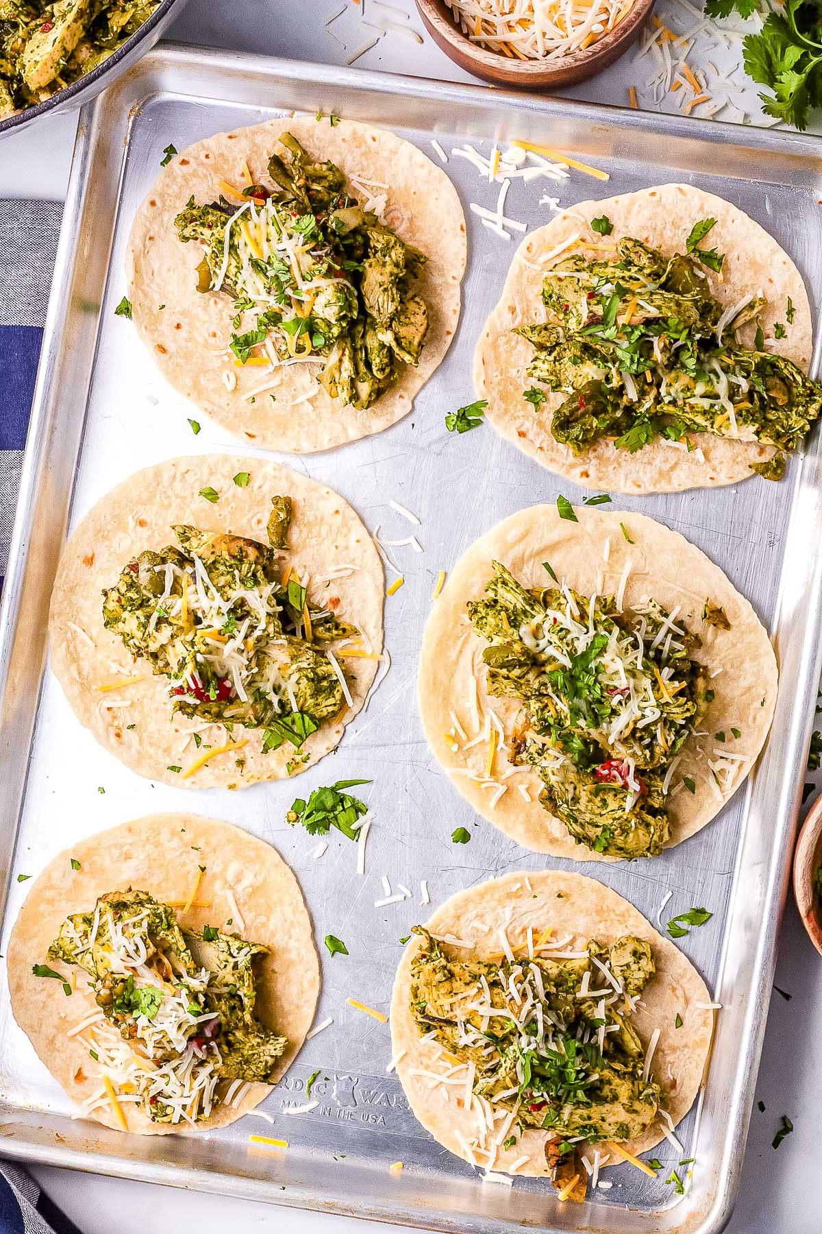 Mini tortillas filled with zhoug chicken taco mixture, shredded cheese, and cilantro on a sheet pan.