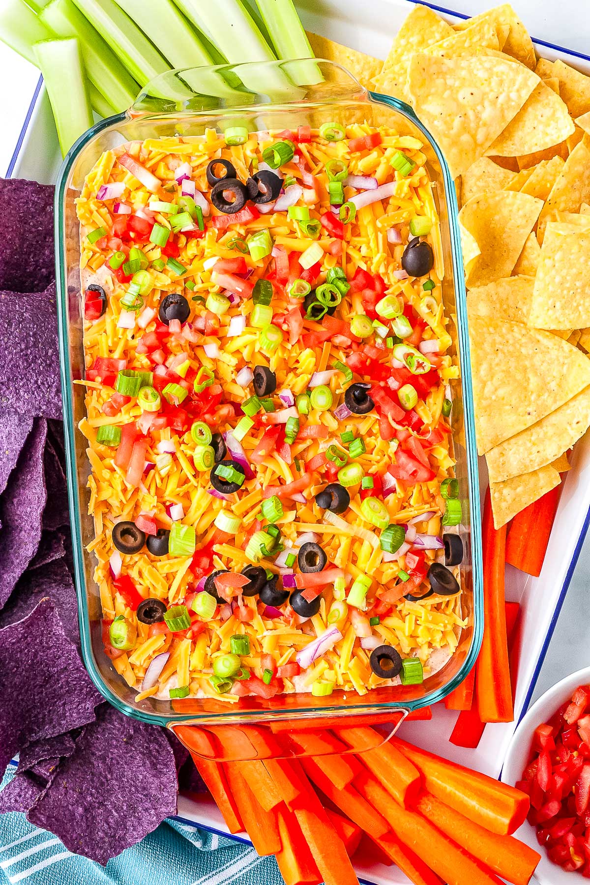 Glass baking dish filled with cottage cheese taco dip, topped with cheddar cheese, tomatoes, green onions, red onions, and black olives, surrounded by tortilla chips and fresh vegetables.