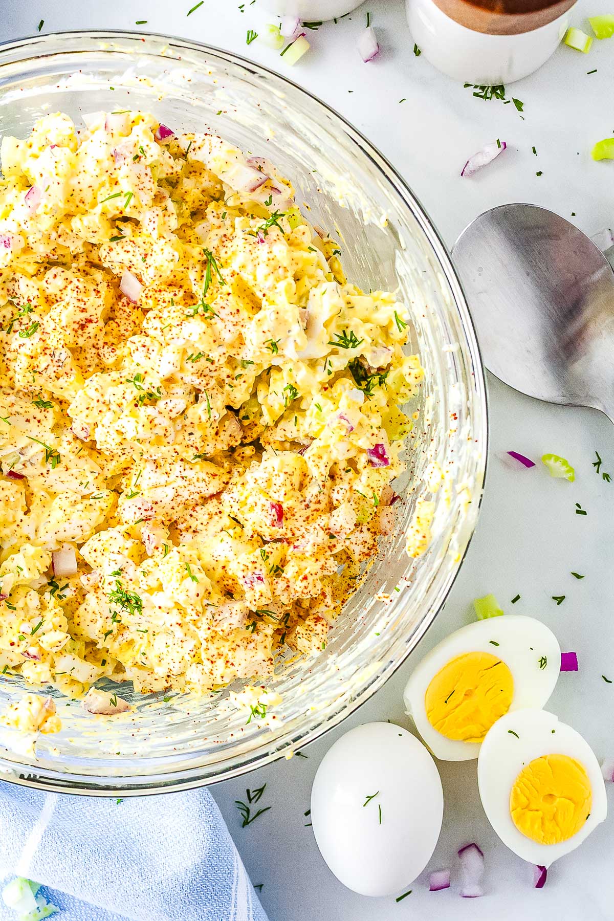 An overhead view of a glass mixing bowl filled with cottage cheese egg salad garnished with paprika and dill. The bowl sits on a white surface next to a silver serving spoon, a blue napkin, and a halved hard-boiled egg.